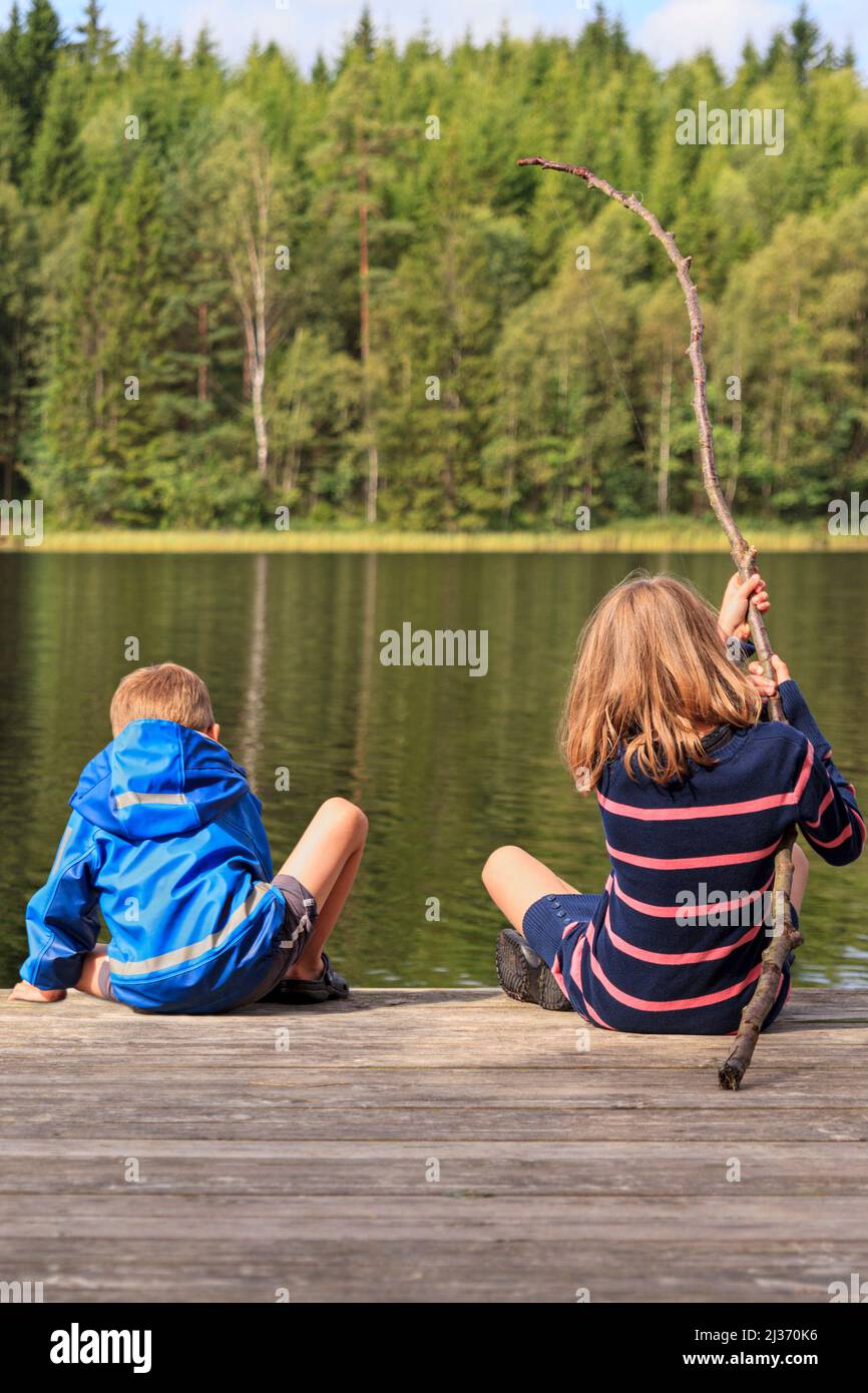 Two children, younger brother and older sister, using a homemade fishing rod fishing from jetty by a lake set in an idyllic Swedish summer forest landscape Stock Photo