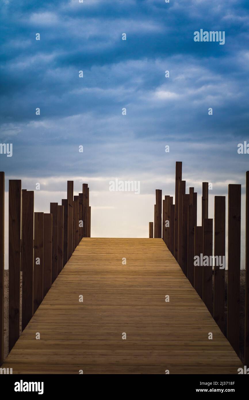 A boardwalk with vertical wood piles on both sides leading to the unknown and a dramatic sky on top. Path to light, hope or future concept Stock Photo
