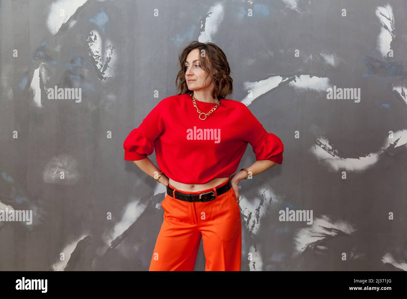 Fashionable and stylish girl posing in orange pants, a red sweater against a gray wall Stock Photo