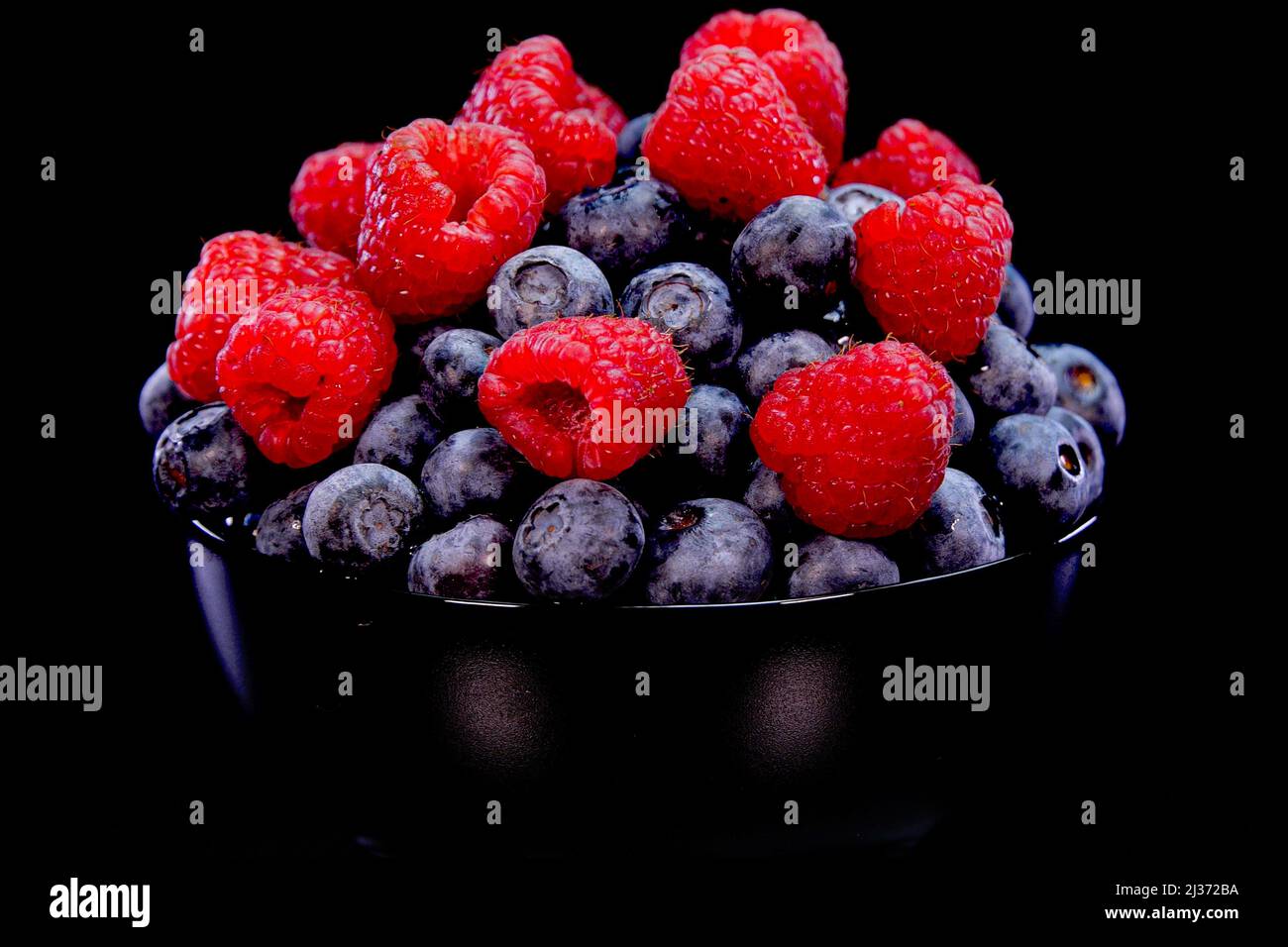 Raspberries and blueberries in a black bowl, on a black background. Fresh, firm and tasty berries. Raw vegan summer snacks, forest berries. Stock Photo