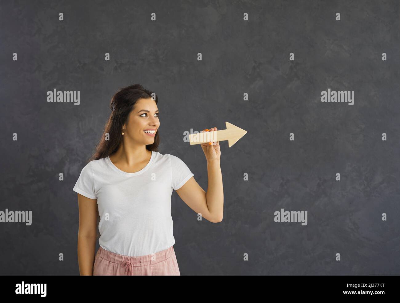 Young caucasian woman standing on a gray background holding a wooden arrow pointing to the side. Stock Photo