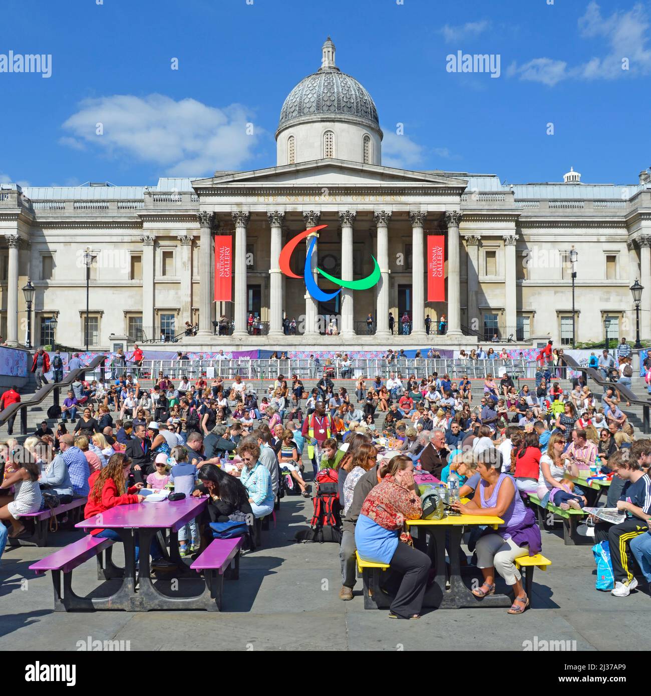 Tourists & visitors sit at coloured picnic tables in Trafalgar Square 2012 London Paralympic Games logo on colonnade of National Gallery England UK Stock Photo