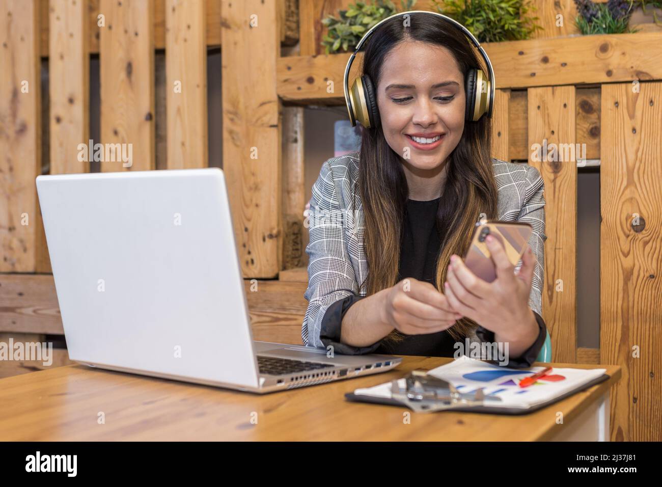 Positive female remote specialist in wireless headphones using cellphone while doing remote job on netbook at table with document on clipboard. Stock Photo