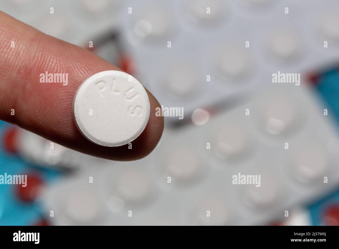 White pill on a finger in selective focus. There is the inscription 'PLUS', which indicates that the drug is more effective. Out of focus drug tablets Stock Photo