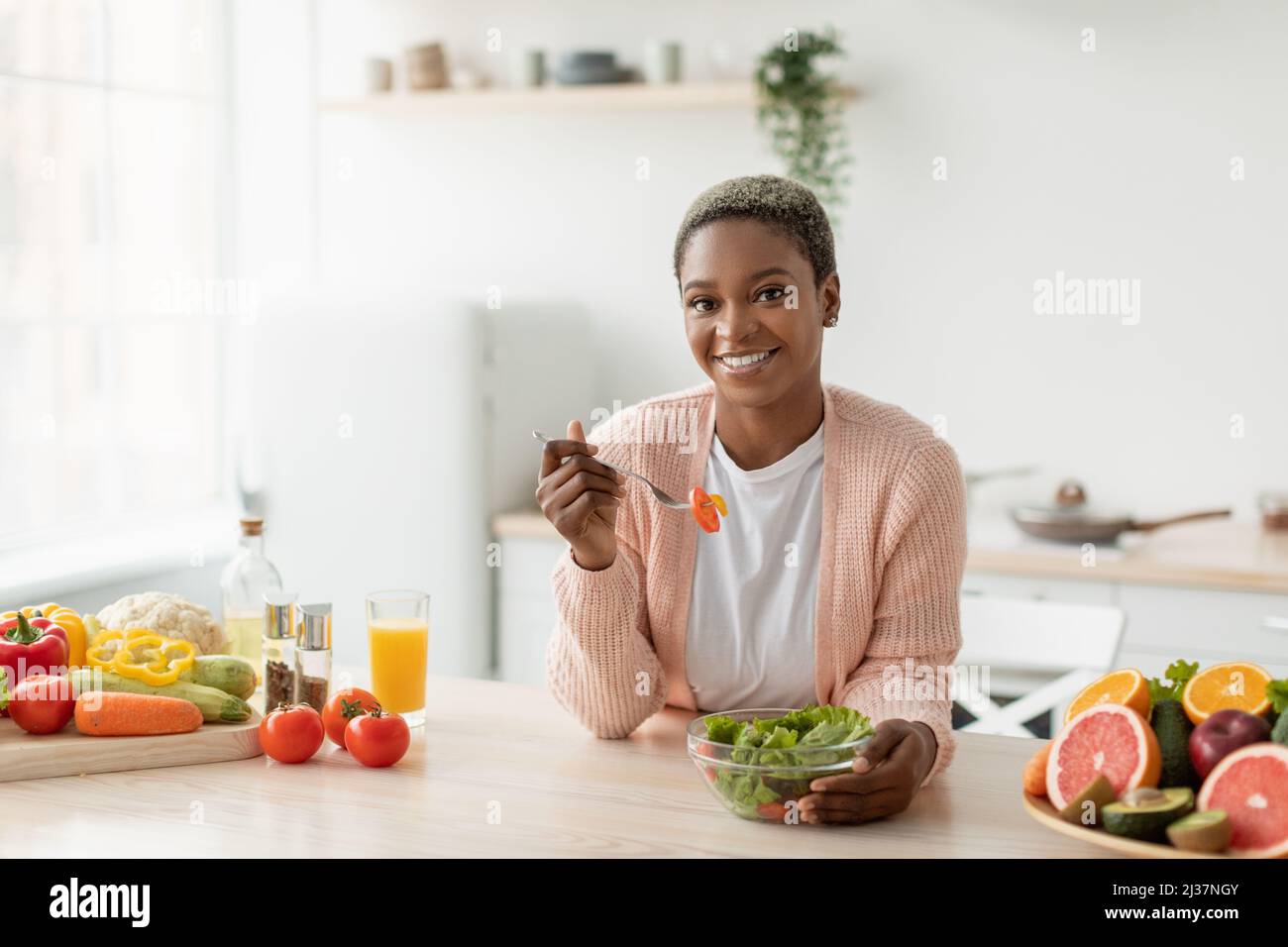 Cheerful millennial african american woman eating salad with organic vegetables in minimalist kitchen interior Stock Photo