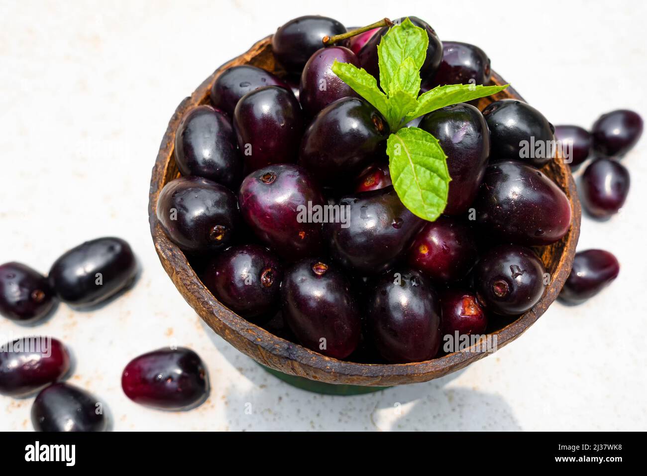 Dark pink-red ripe Syzygium cumini fruits. Dark black java plum in a wood bowl at isolated white background. Green mint leaf on top of some large Stock Photo