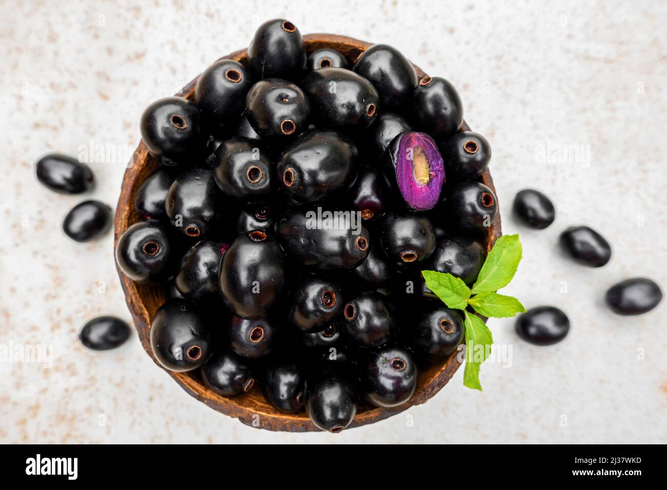 Black ripe Syzygium cumini fruits. Dark black java plum in a wood bowl at isolated white background. Green mint leaf on top of some large java plums. Stock Photo