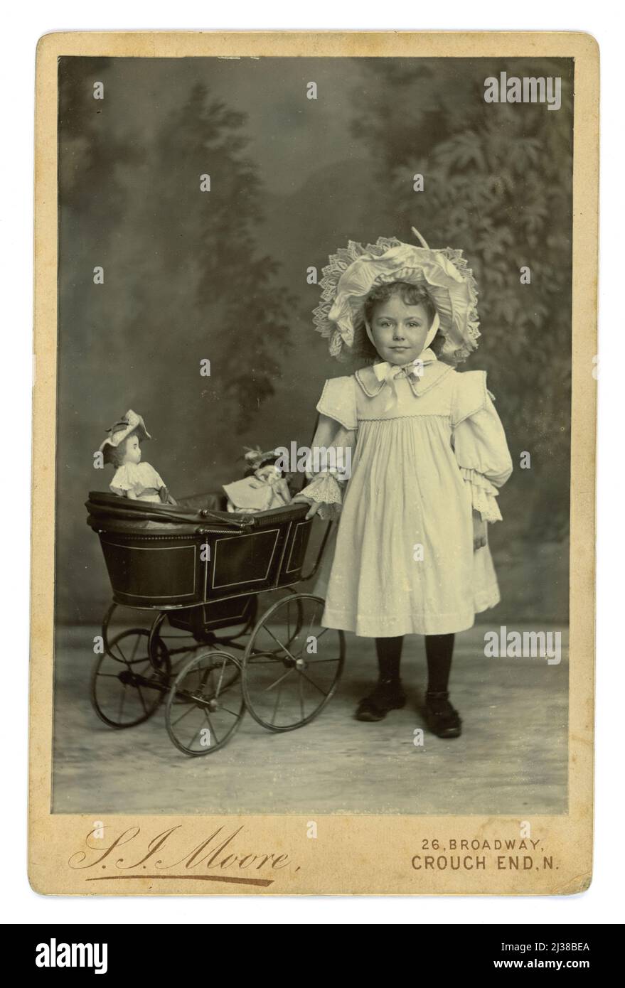 Original, very clear, Edwardian era cabinet card of adorable cute young Edwardian or Victorian girl from a wealthy middle class London family, standing with her dolls pram, wearing a white bonnet and frock, from the studio of  S. J. Moore 26 Broadway, Crouch End, N. London, circa 1904 Stock Photo
