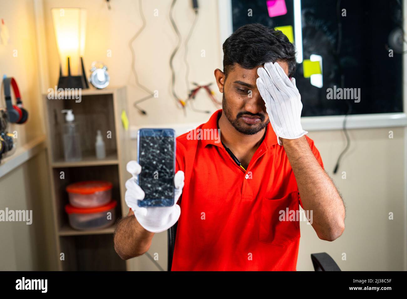 Worried repairman showing broken mobile phone with hands on forehead at workshop - concept of mistakes, skill worker and professional occupation Stock Photo