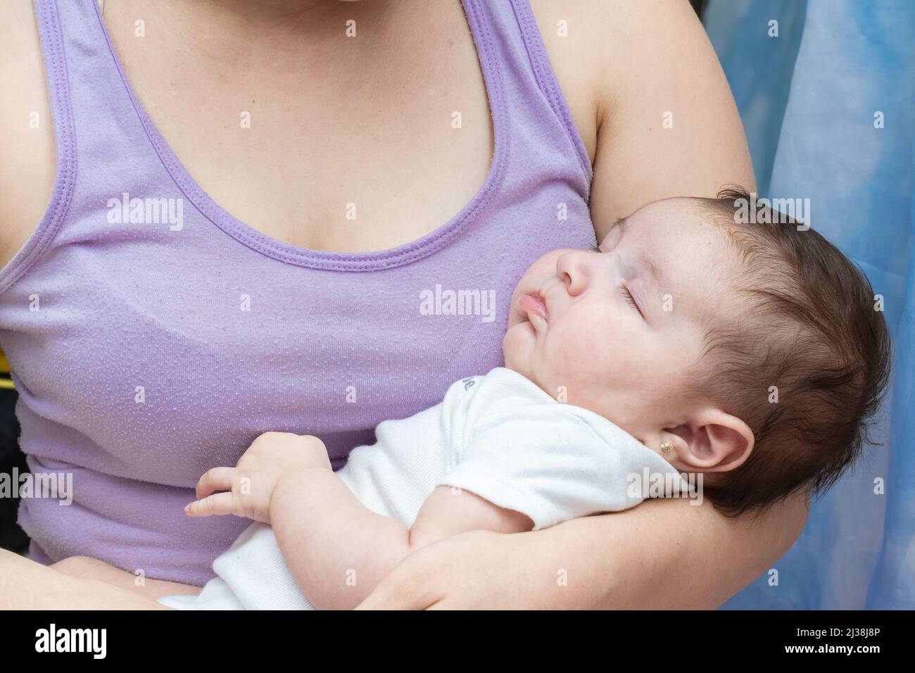beautiful two month old latina baby girl sleeping on her mother's arms. cute baby girl dressed in a white jumpsuit clinging to her mother's chest as s Stock Photo