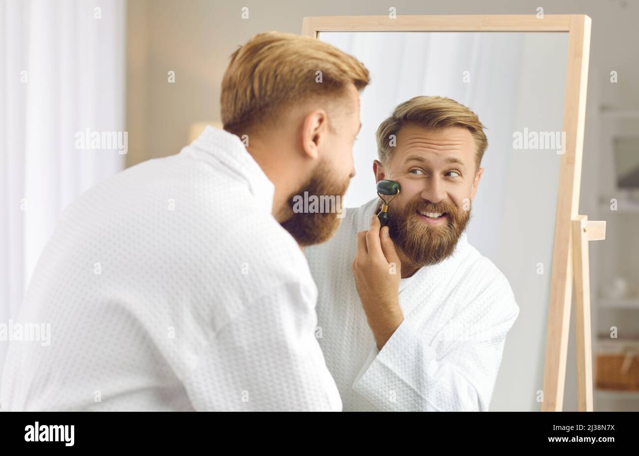 Happy man looking in mirror and massaging his face with jade facial beauty roller Stock Photo