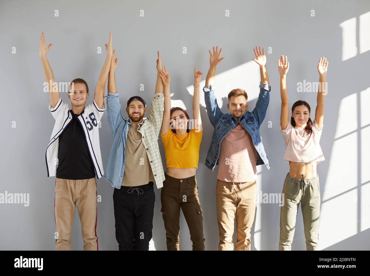 Team of cheerful young people standing together, smiling and raising their hands up Stock Photo