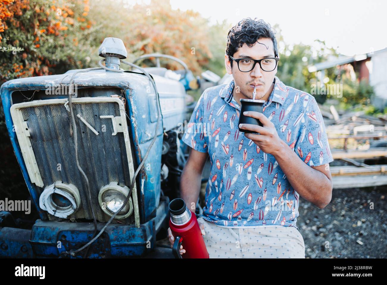 Young latin man drinking mate in a natural space, beside an old little blue tractor. Latin beverage. Stock Photo