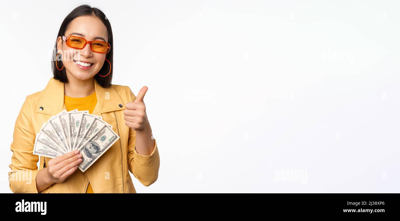 Stylish smiling asian girl holding money cash, showing dollars and celebrating, standing over white background Stock Photo