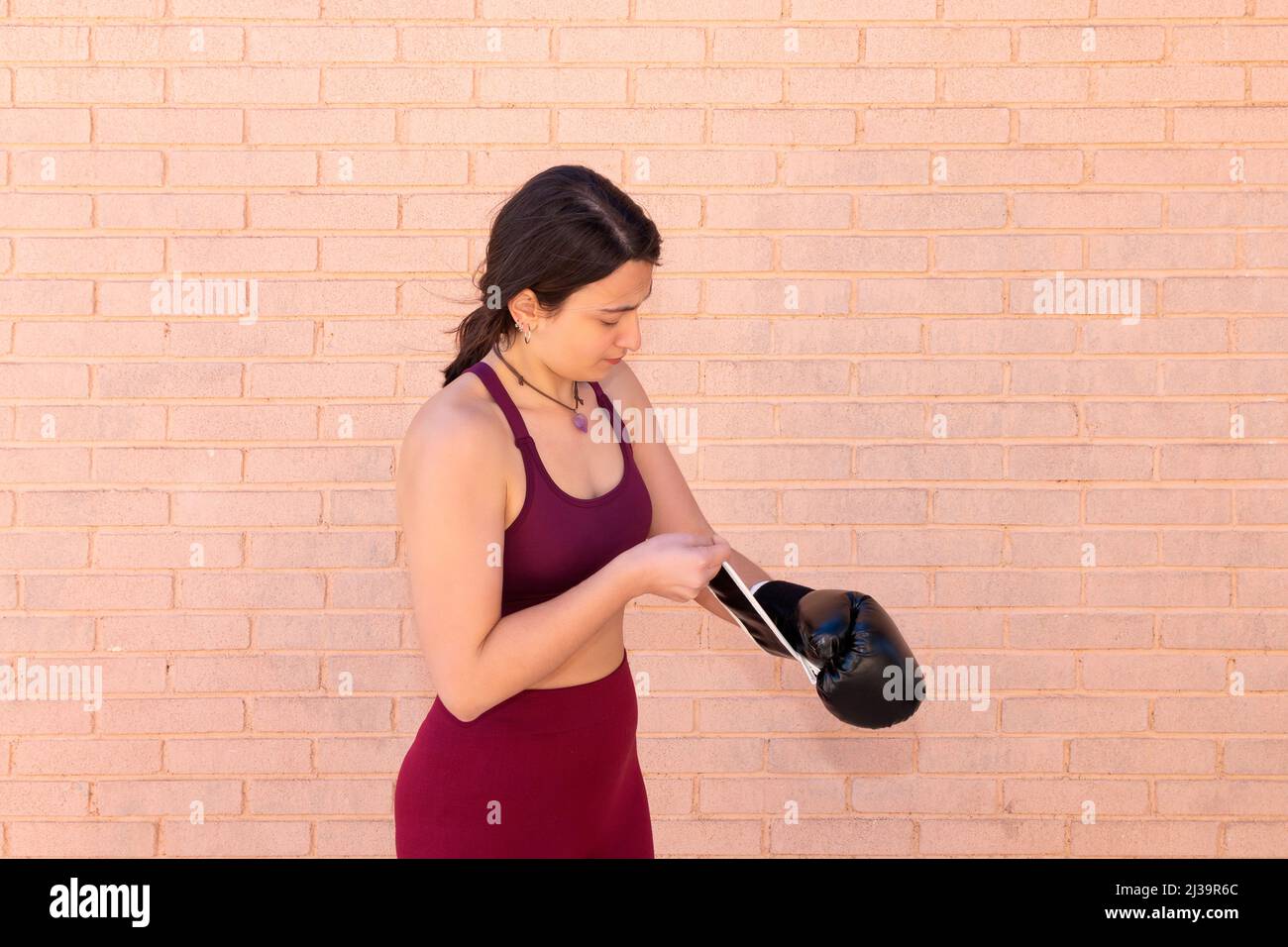 A young Caucasian woman dressed in a top is putting on a black boxing glove in front of a brick wall. Stock Photo
