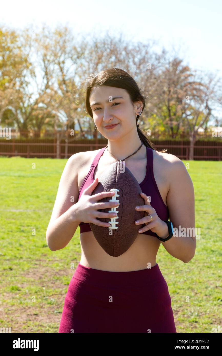 A young Caucasian woman is holding an American football ball with both hands, grasping it by the strings. The person is on the grass of a playing fiel Stock Photo