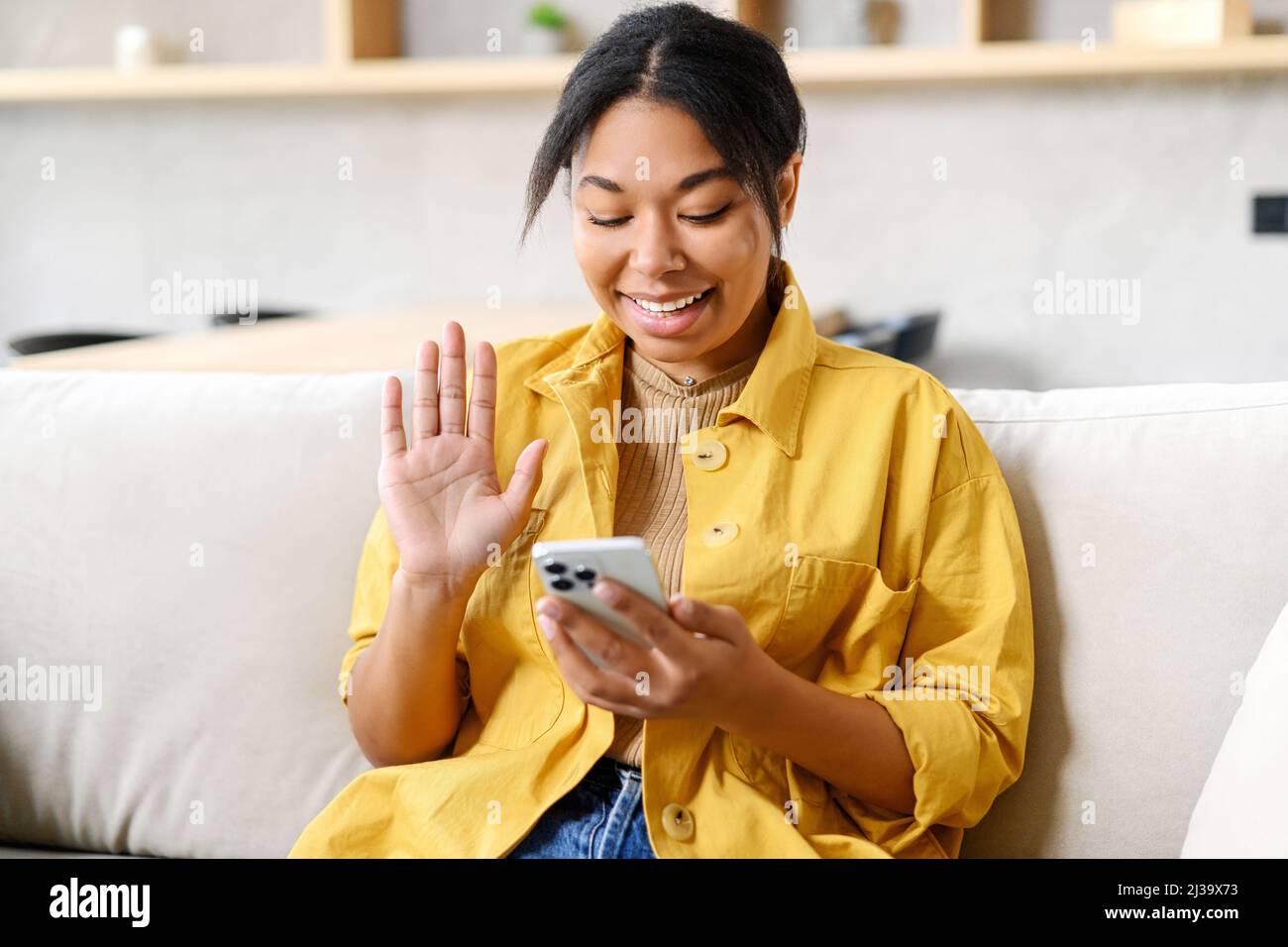 Happy African-American female using mobile app on smartphone for making video call, waving hand and greeting friends or family, talking via video connection, stay in touch on the distance Stock Photo
