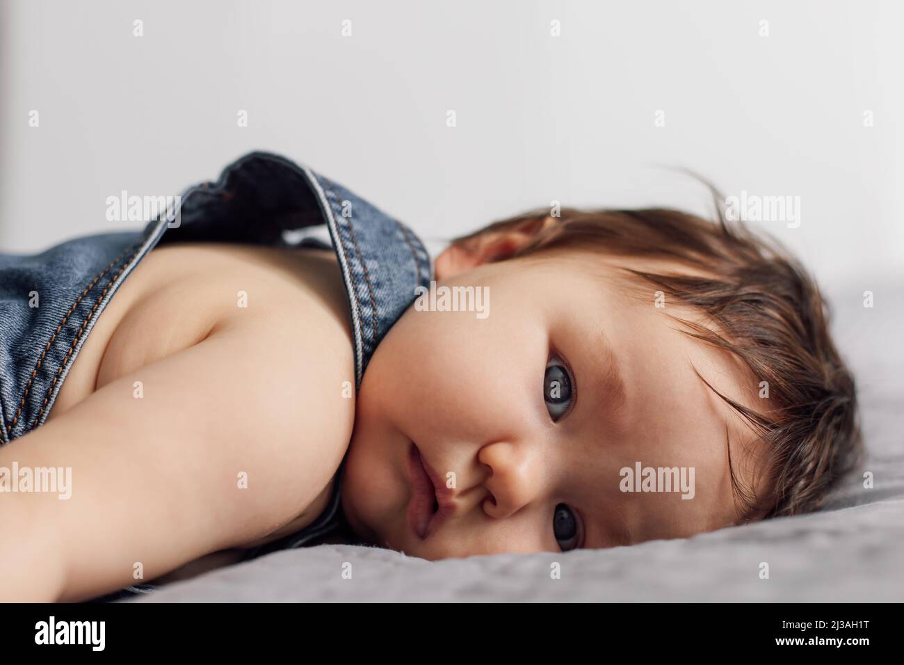 Portrait of pretty baby wearing denim romper lying on gray blanket at home. Tired blue eyed infant child rest on bed in bedroom, white background Stock Photo