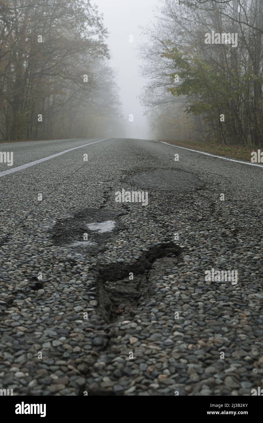 Potholes on a paved road. Nearby you can see traces of repairing another pothole. Bad condition of the country road. Broken road in the foggy forest Stock Photo