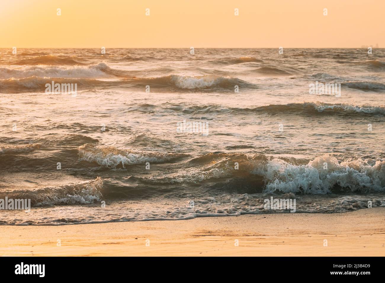 Sea ocean water surface with foaming small waves at sunset. Evening sunlight sunshine above sea. Ocean water foam splashes washing sandy beach Stock Photo