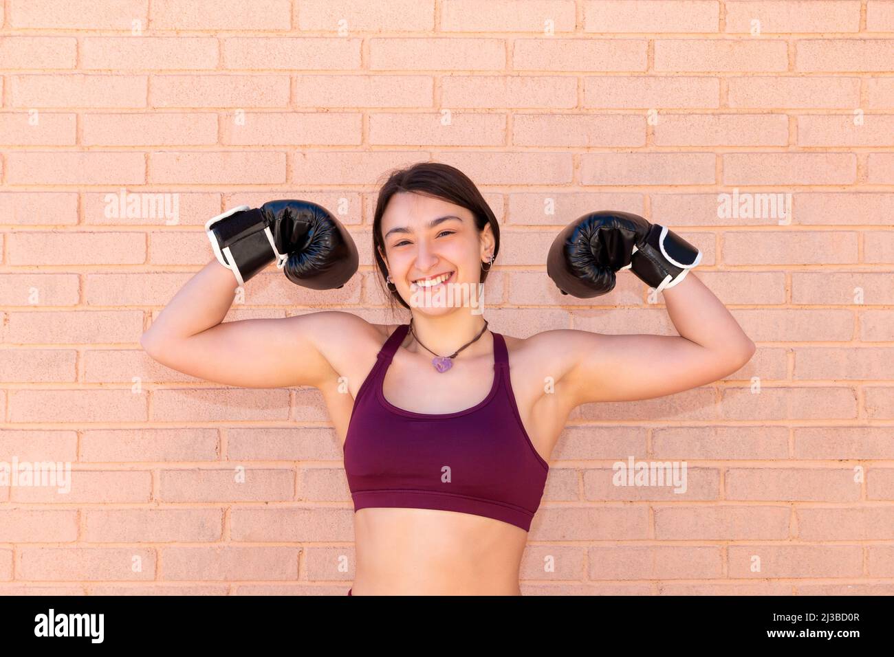 A smiling young Caucasian woman wearing black boxing gloves exerting force with her arms showing off her biceps. In the background is a brick wall. Stock Photo