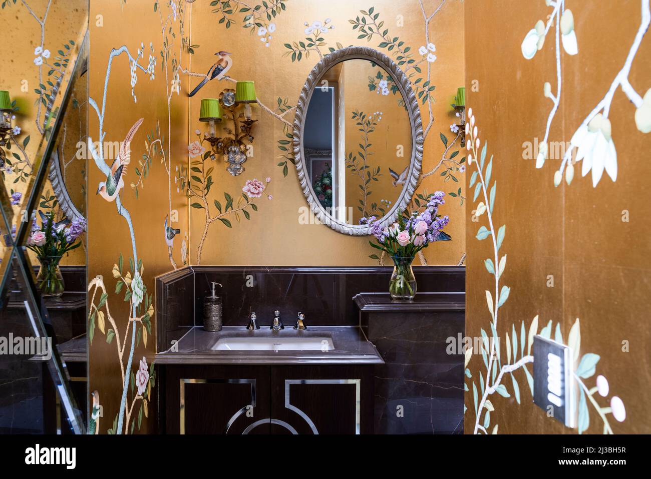 Gold Chinoiserie de Gournay  wallpaper with oval mirror above washbasin in Holland Park home, London, UK Stock Photo