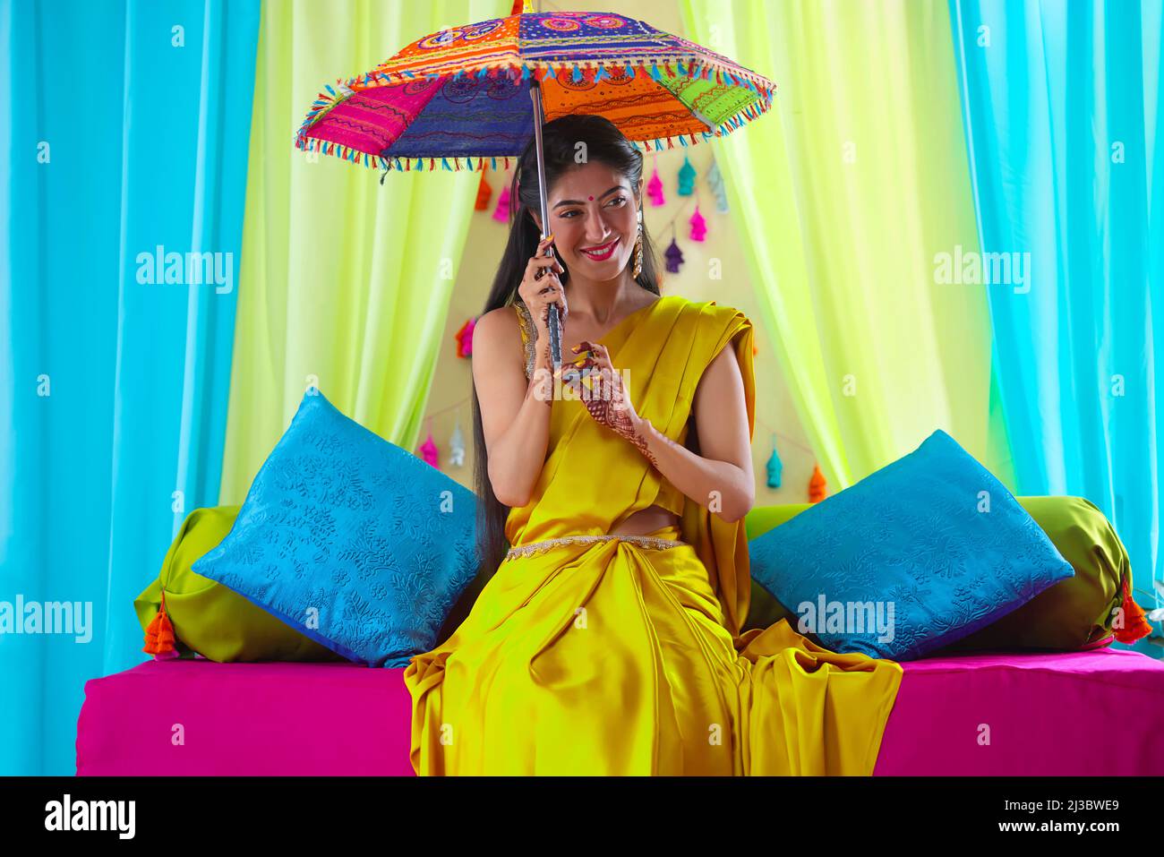 Indian bride in yellow saree sitting on stage with holding umbrella over head during haldi ceremony Stock Photo