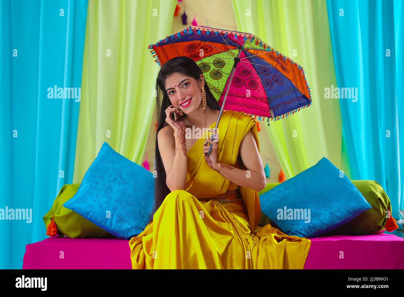 Indian bride in yellow saree looking at camera while sitting on stage with holding an umbrella over head Stock Photo
