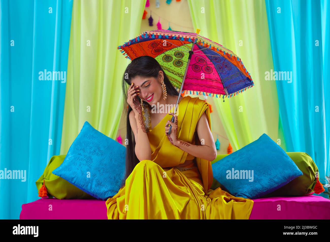 Indian bride in yellow saree looking down while sitting on stage with holding a umbrella over head Stock Photo