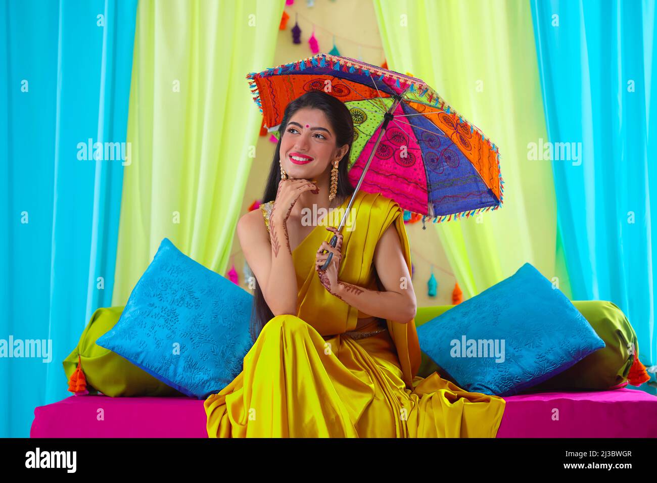Indian bride in yellow saree looking elsewhere while sitting on stage with holding a umbrella over head Stock Photo
