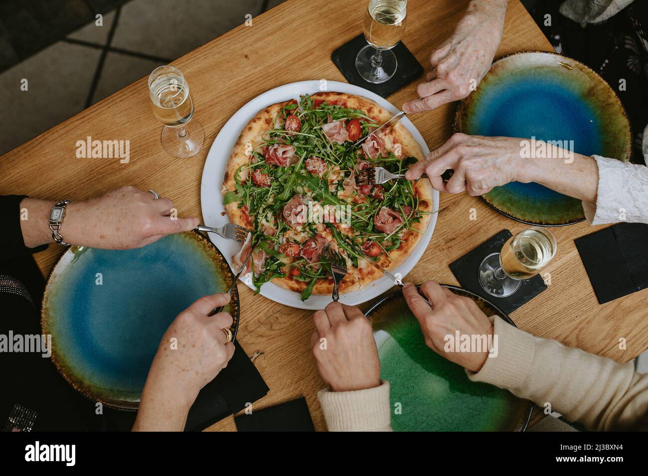 Top view of unrecognizable senior women sitting around wooden table with glasses of champagne, digging forks into pizza. Stock Photo