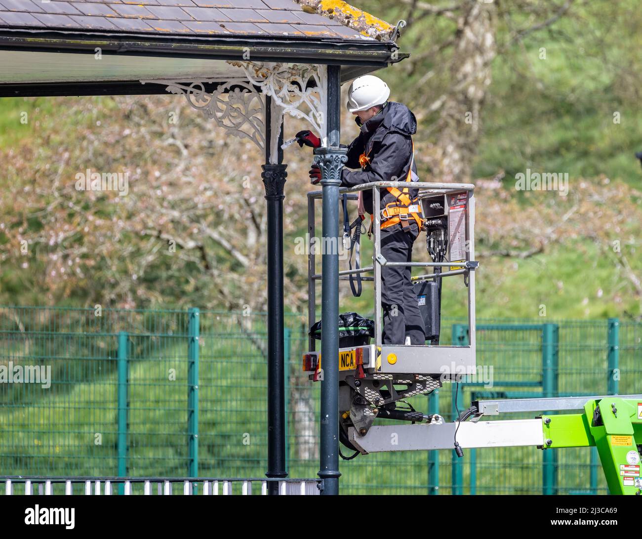Workman on cherry picker with safety harness painting band stand metalwork in Warminster, Wiltshire, UK on 7 April 2022 Stock Photo