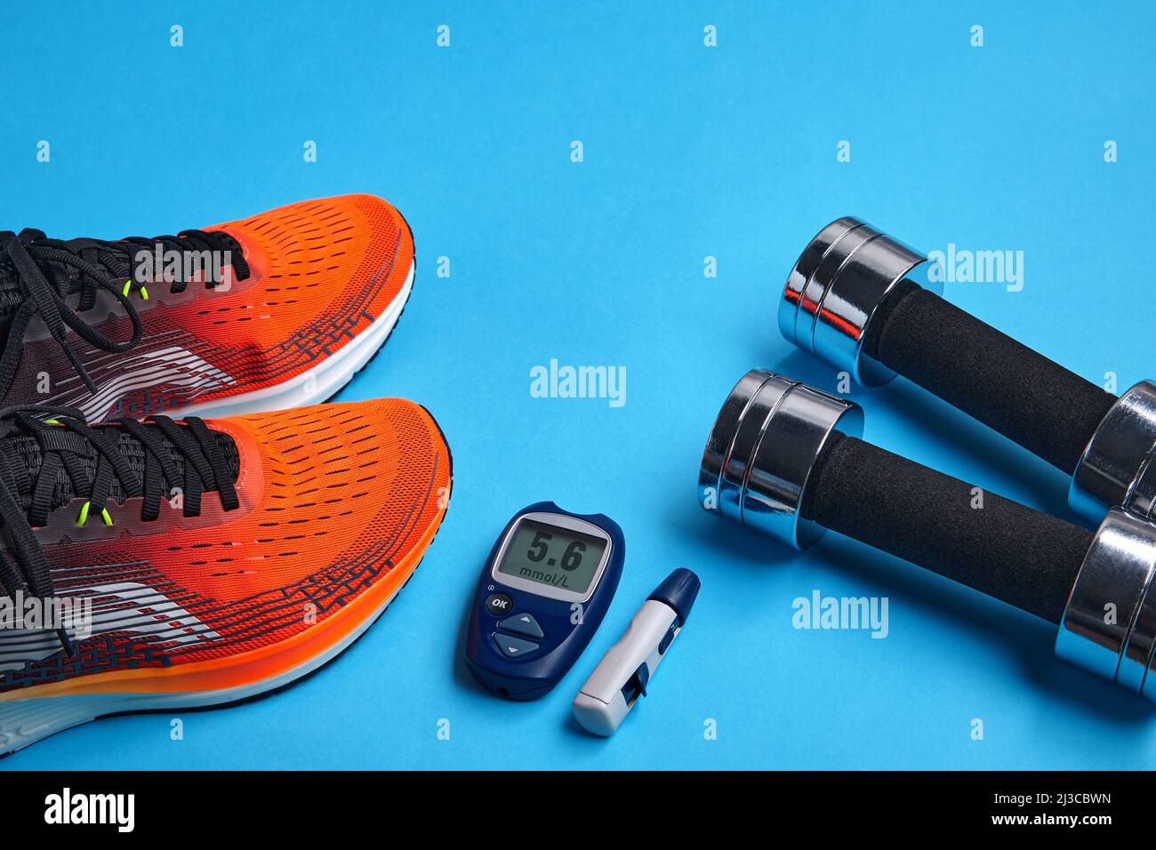 Orange sneakers, metal dumbbells and a blood glucose meter on a blue background. Exercise to overcome insulin resistance Stock Photo