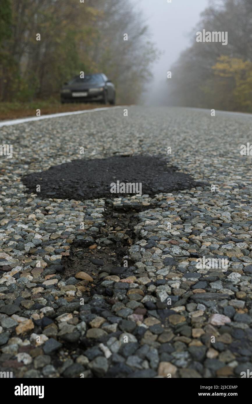 Potholes and a patch on a paved road that runs through a foggy forest. There is a car standing on the side of the road ahead. The poor condition of th Stock Photo