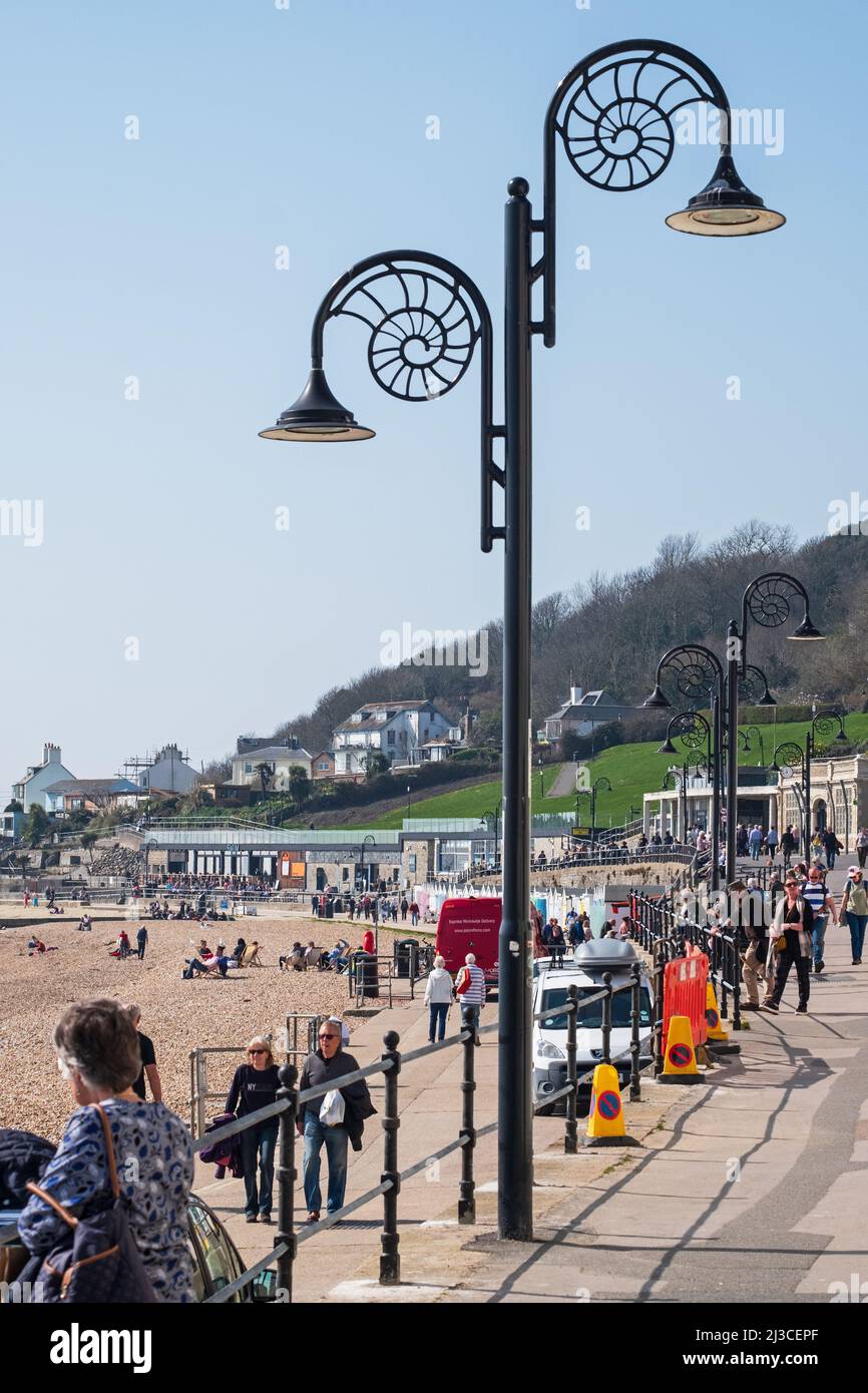 People on the Lyme Regis seafront beneath the ammonite inspired lampposts on a sunny Spring day in the resort on the Jurassic coast famous for fossils Stock Photo