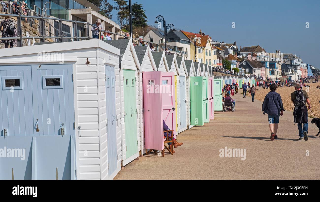 People strolling past traditional Victorian beach huts lining the seafront on a sunny Spring day in the Dorset resort of Lyme Regis UK Stock Photo