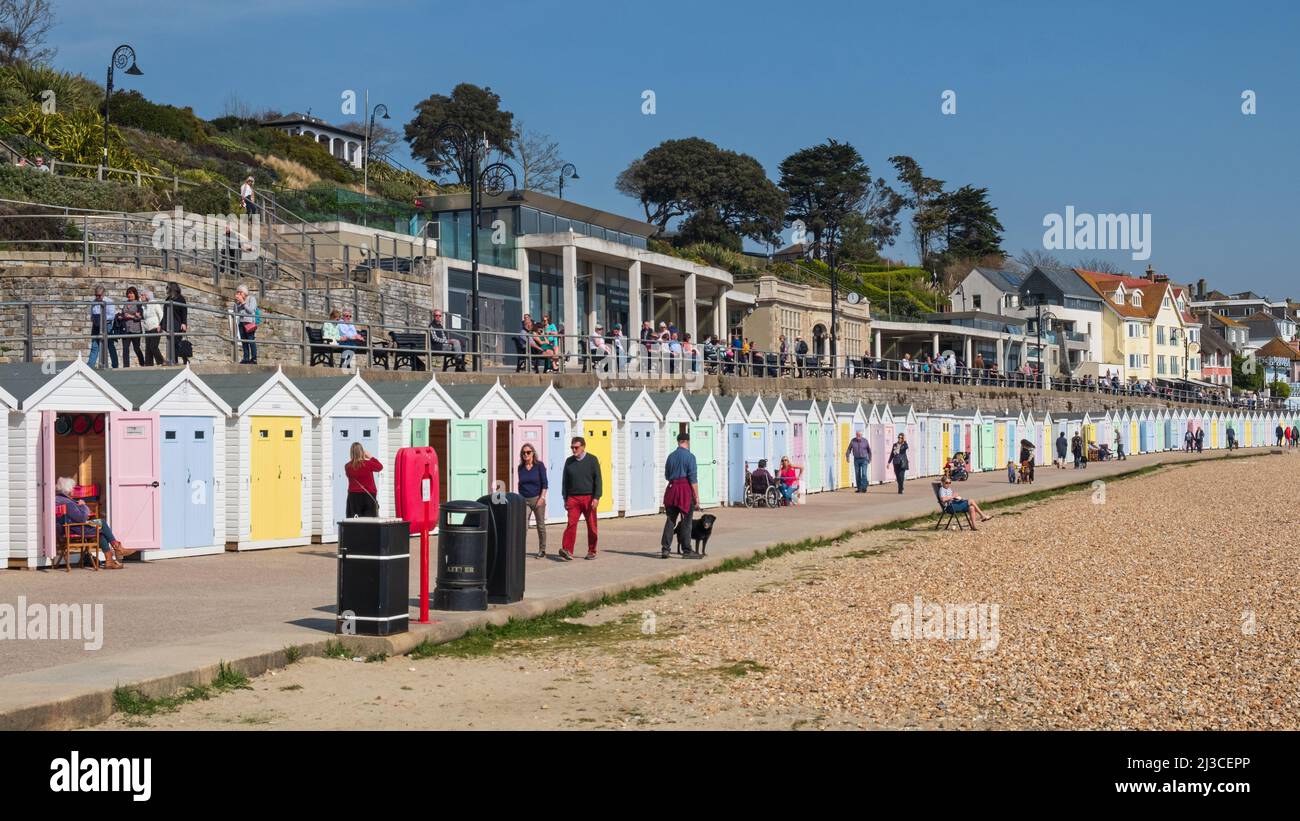People strolling past traditional Victorian beach huts lining the seafront at Lyme Regis on a sunny Spring day in the Dorset resort UK Stock Photo