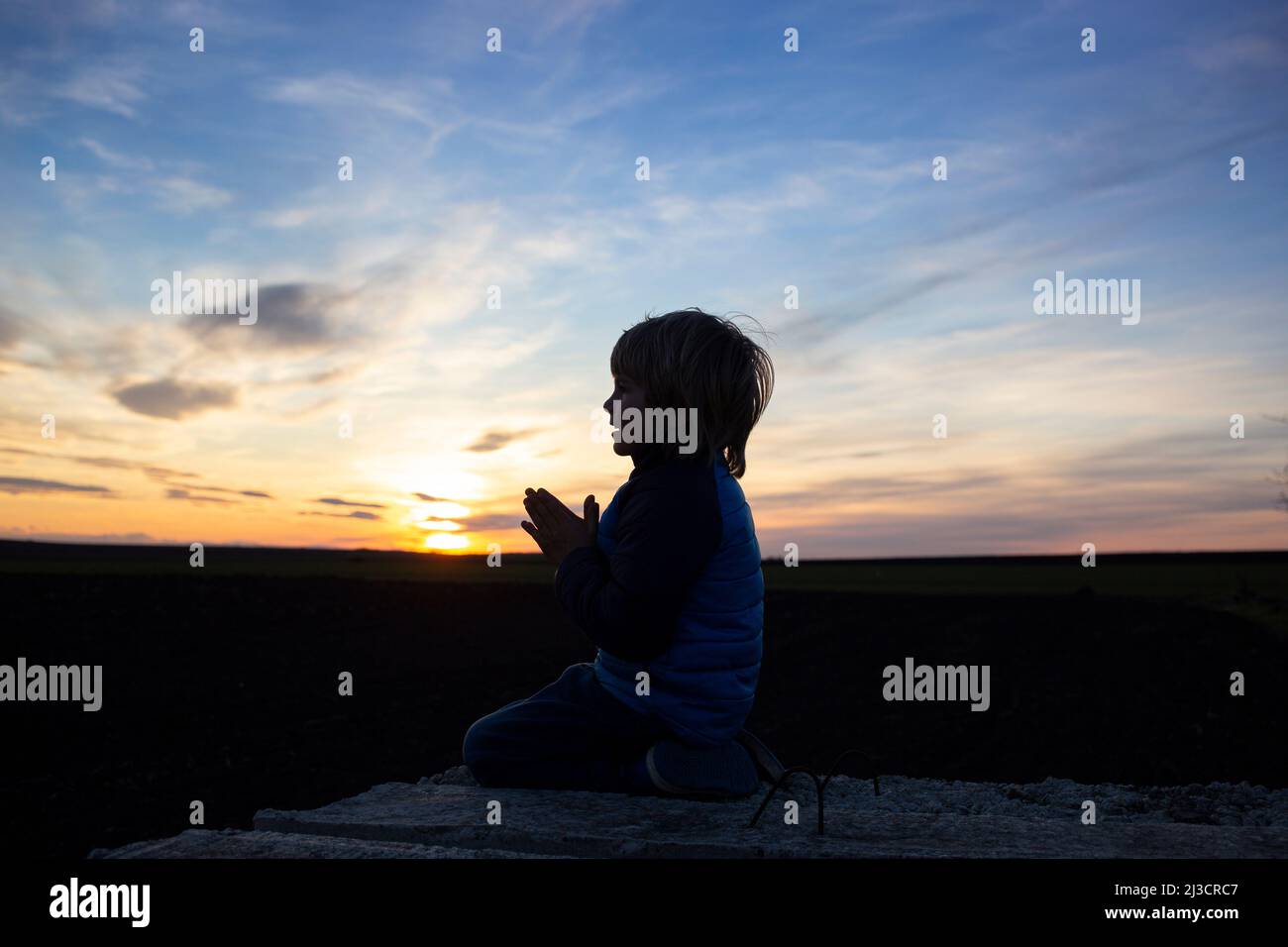 silhouette of a boy sitting on his knees, hands folded in prayer, turning to God at sunset. Children against war. the child asks for peace for his hom Stock Photo