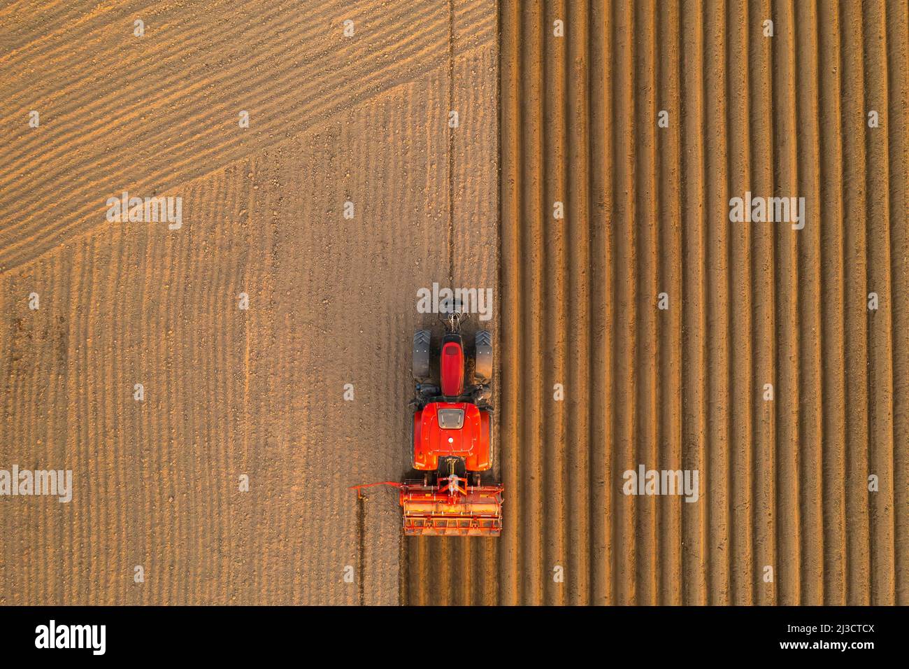 PRAGUE , CZECH REPUBLIC - MARCH 18 2022: Agricultural machine prepares long beds for planting potatoes on cultivated field. Red tractor works dragging plow behind aerial view Stock Photo