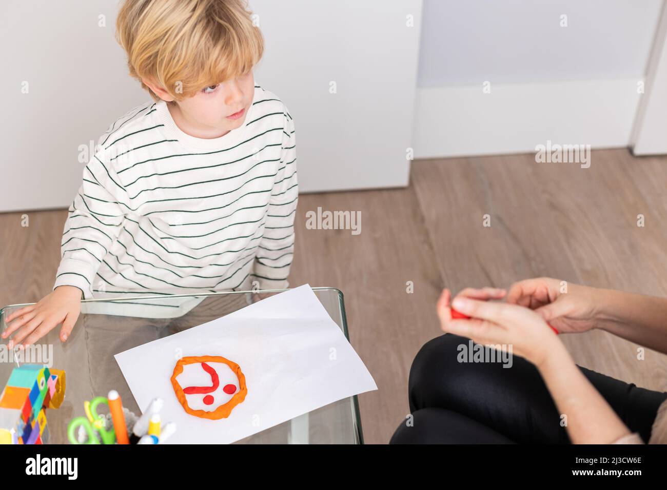 Cute boy sitting at table with sad face on paper made of plasticine near crop unrecognizable psychologist during appointment Stock Photo