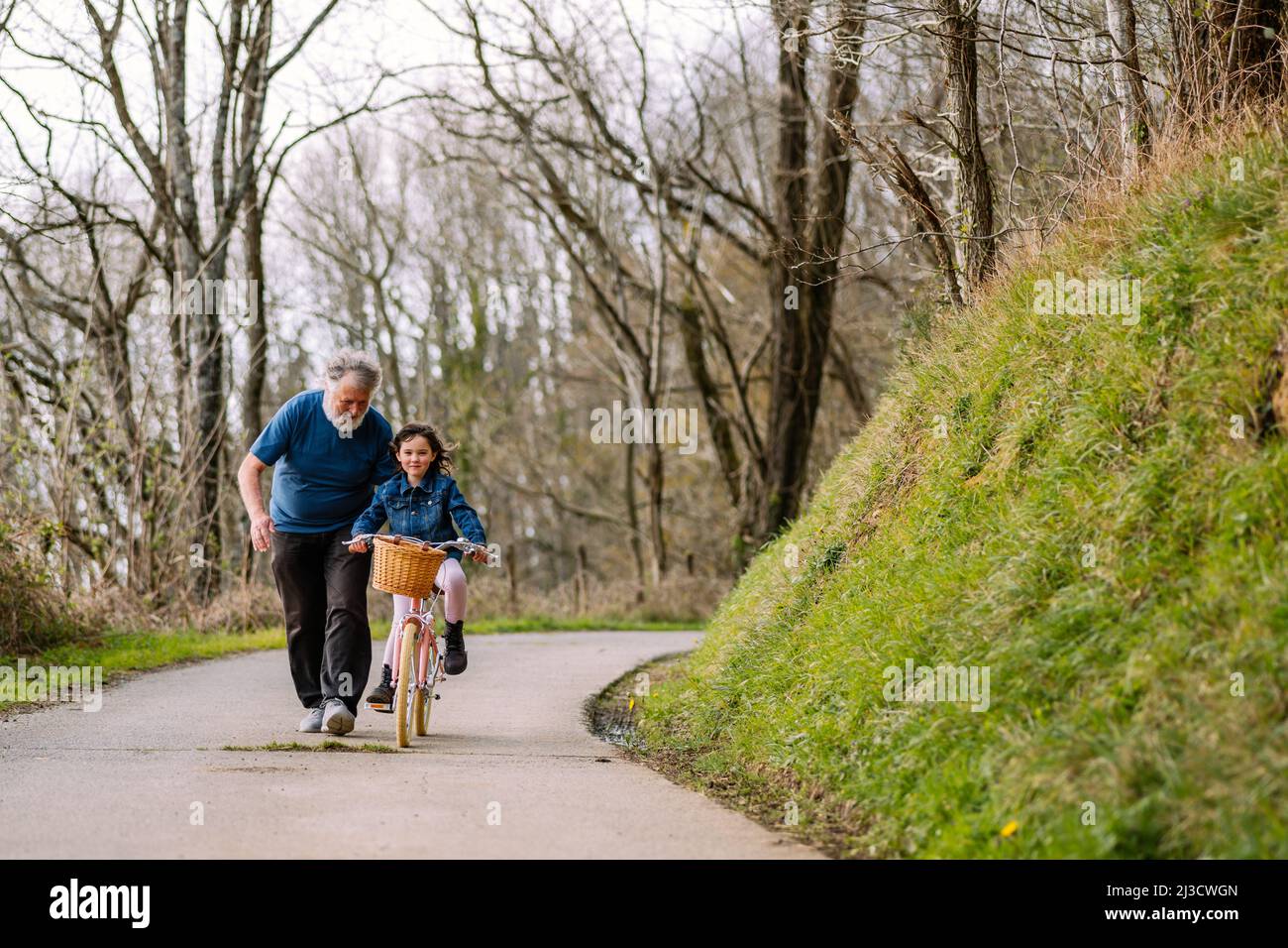 Caring grandfather helping active granddaughter riding bicycle with basket on road in countryside with green trees on summer day Stock Photo