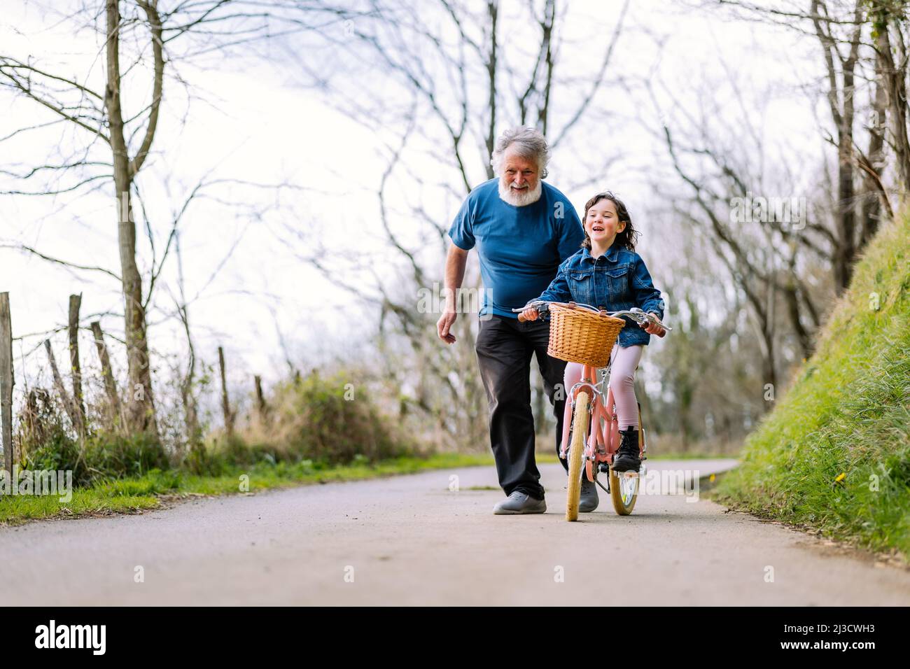 Distant caring grandfather helping active granddaughter riding bicycle with basket on road in countryside with green trees on summer day Stock Photo