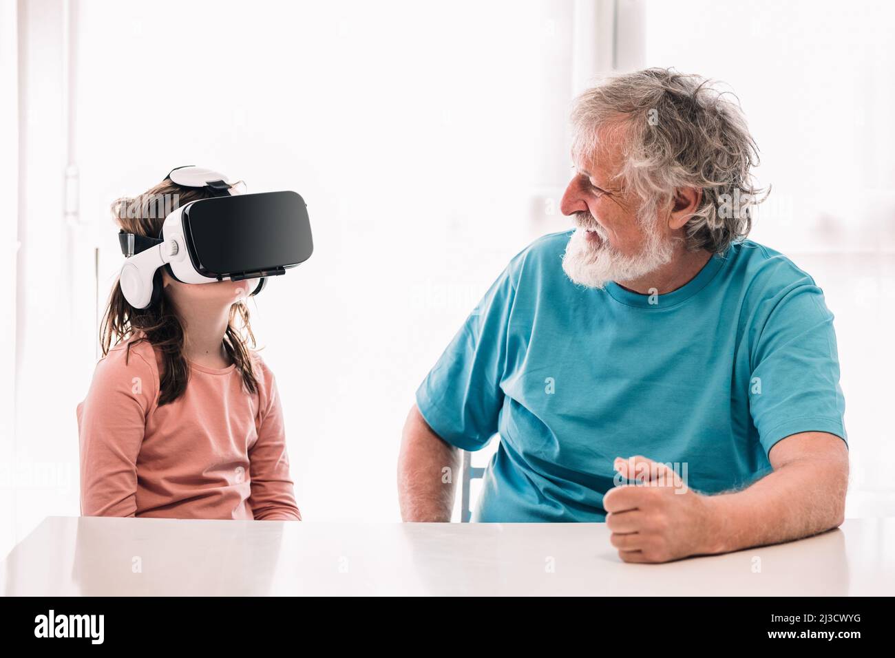 Cute girl in modern VR goggles exploring cyberspace while sitting near bearded grandfather in light room at home Stock Photo