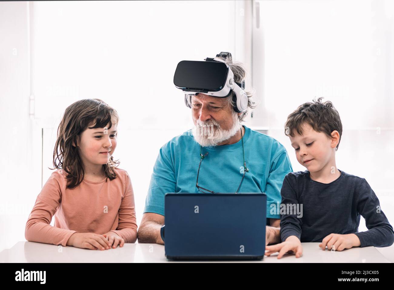 Bearded grandfather in modern VR headset playing videogame on laptop while sitting at table near cute children in light room Stock Photo