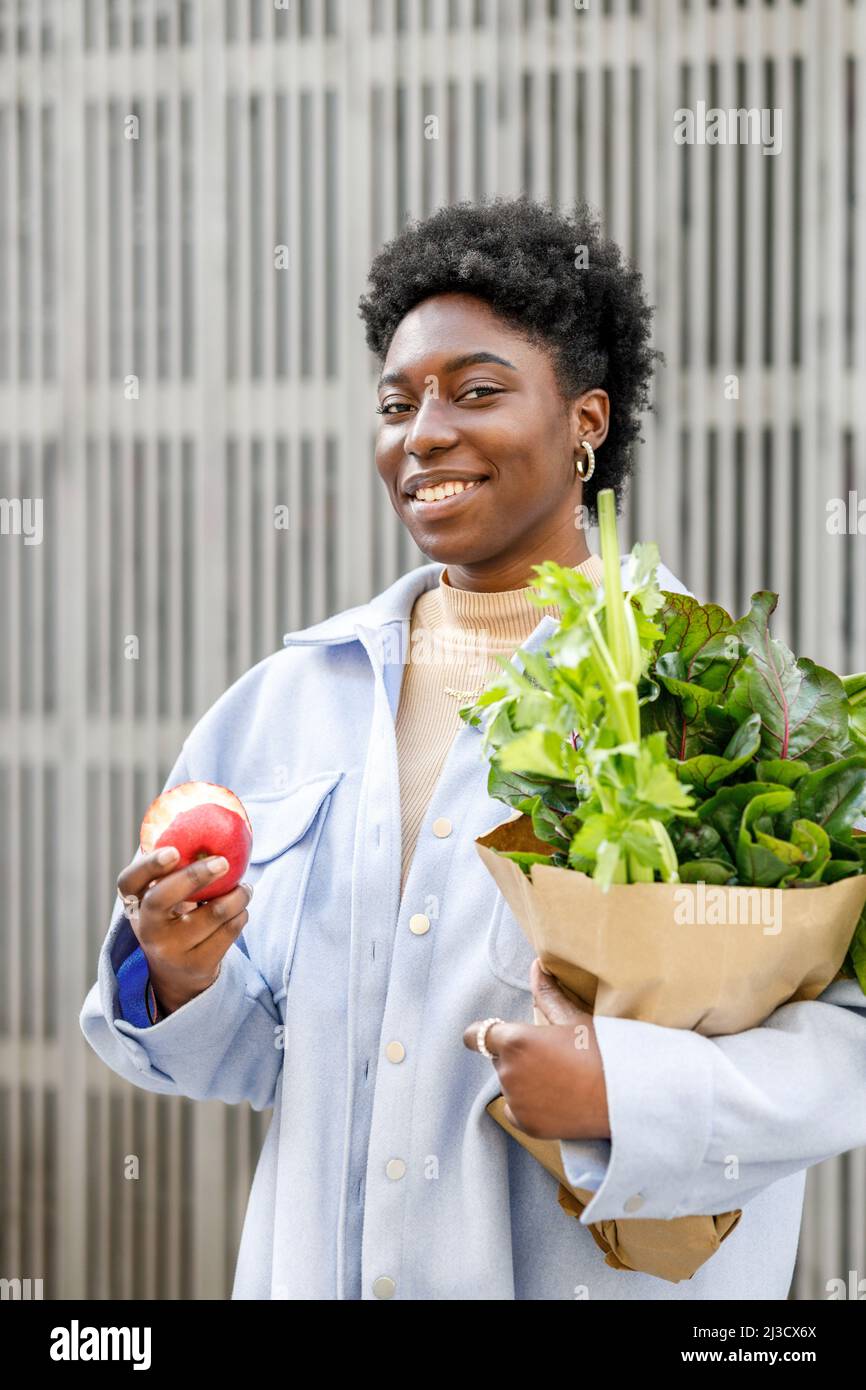 Positive African American female with paper bag of greens eating apple
