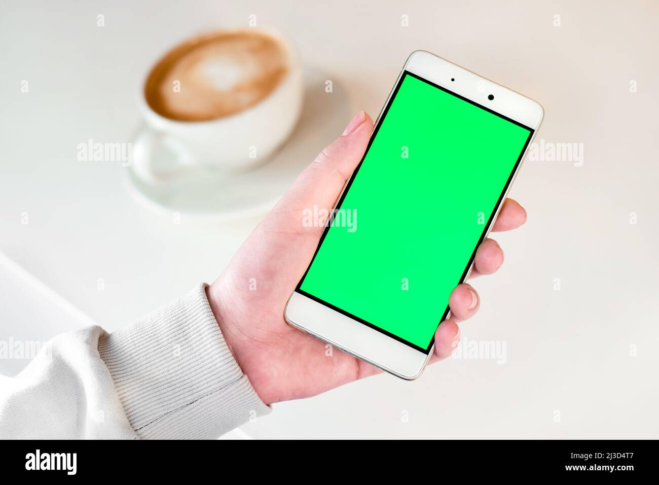Hand holding green Screen Phone with a Cup of Cappuccino on white table. Woman using Mobile Phone with Chroma Key. Over shoulder View. Stock Photo