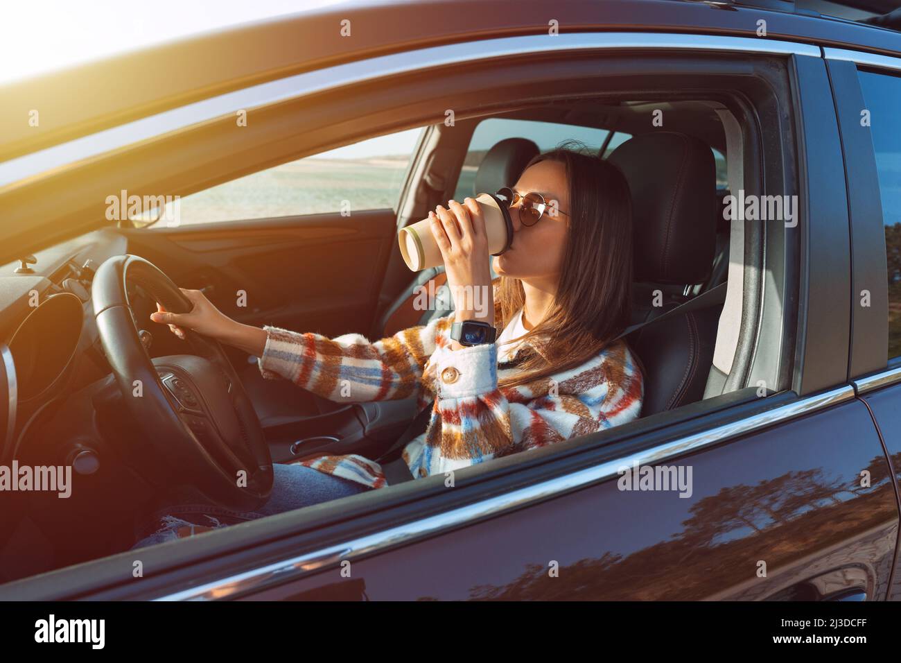 Young beautiful stylish woman driving her new car and drinking coffee on a sunny day. Concept for car rental Stock Photo