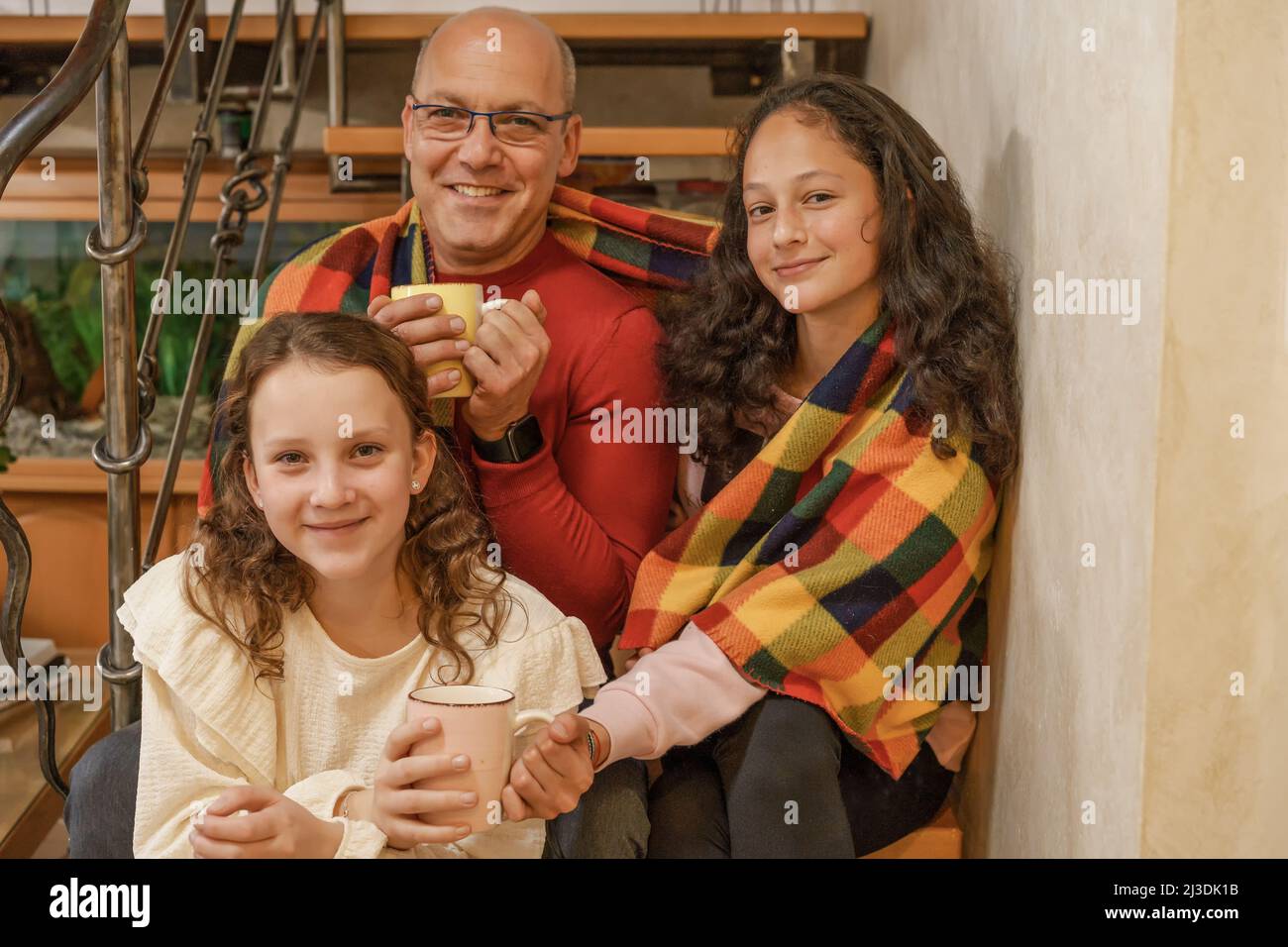 Father and two teenage daughters sitting in the wood stairs wrapped in blanket with a cup of hot drink. Cold in home, problems with household heat supply in winter. Stock Photo