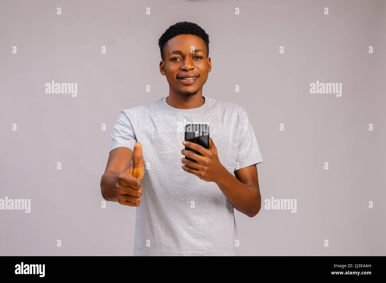cute guy isolated over grey background smiling as he holds his phone and did thumbs up Stock Photo