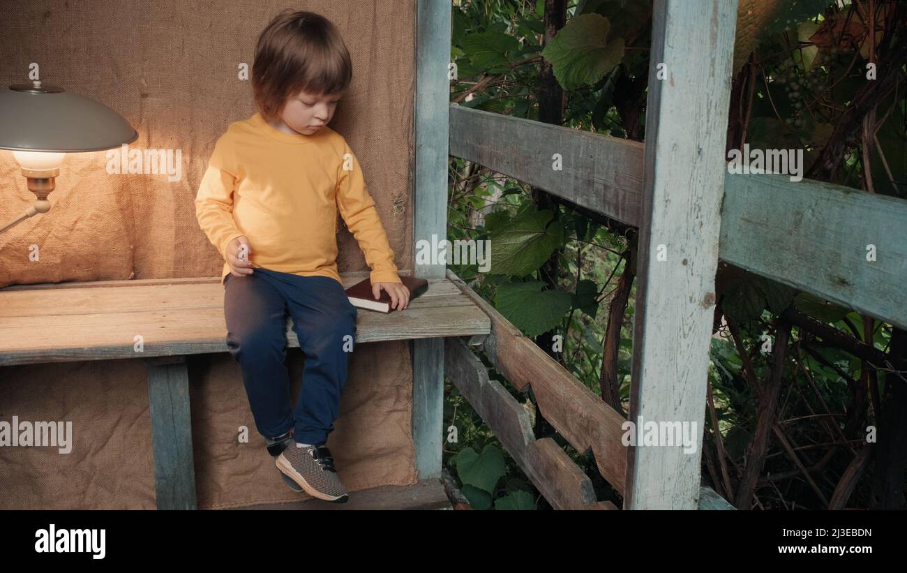 Cute little girl on the porch of a village house is trying to read a book Stock Photo